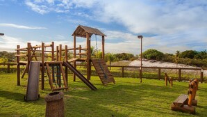 Un’area giochi in legno con scivoli e elementi di arrampicata, circondata da un prato recintato e con una spiaggia sullo sfondo.