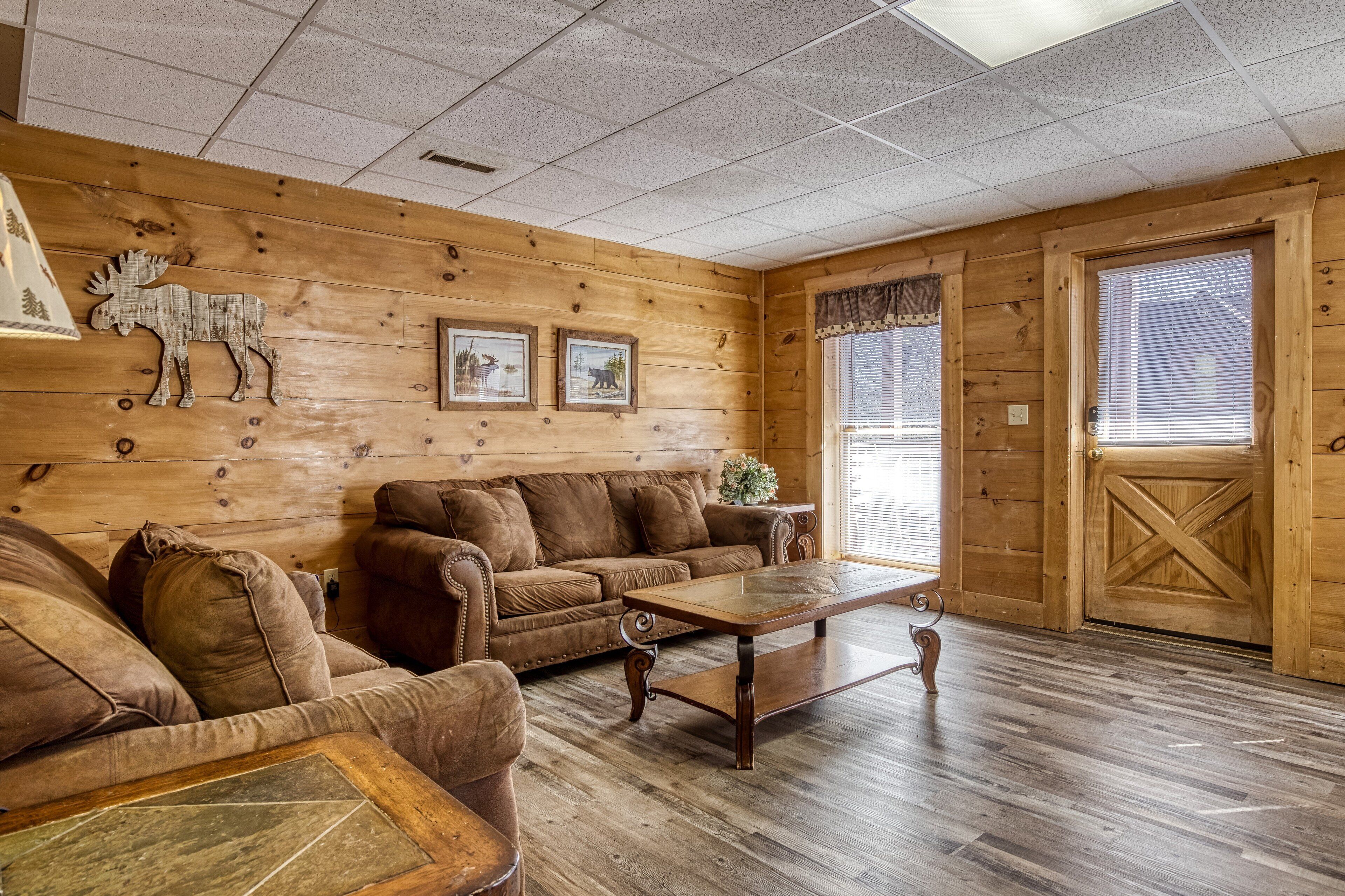 A cozy log cabin interior with a wooden floor, a brown leather sofa, a wooden coffee table, and a wooden door with a cross-brace design.