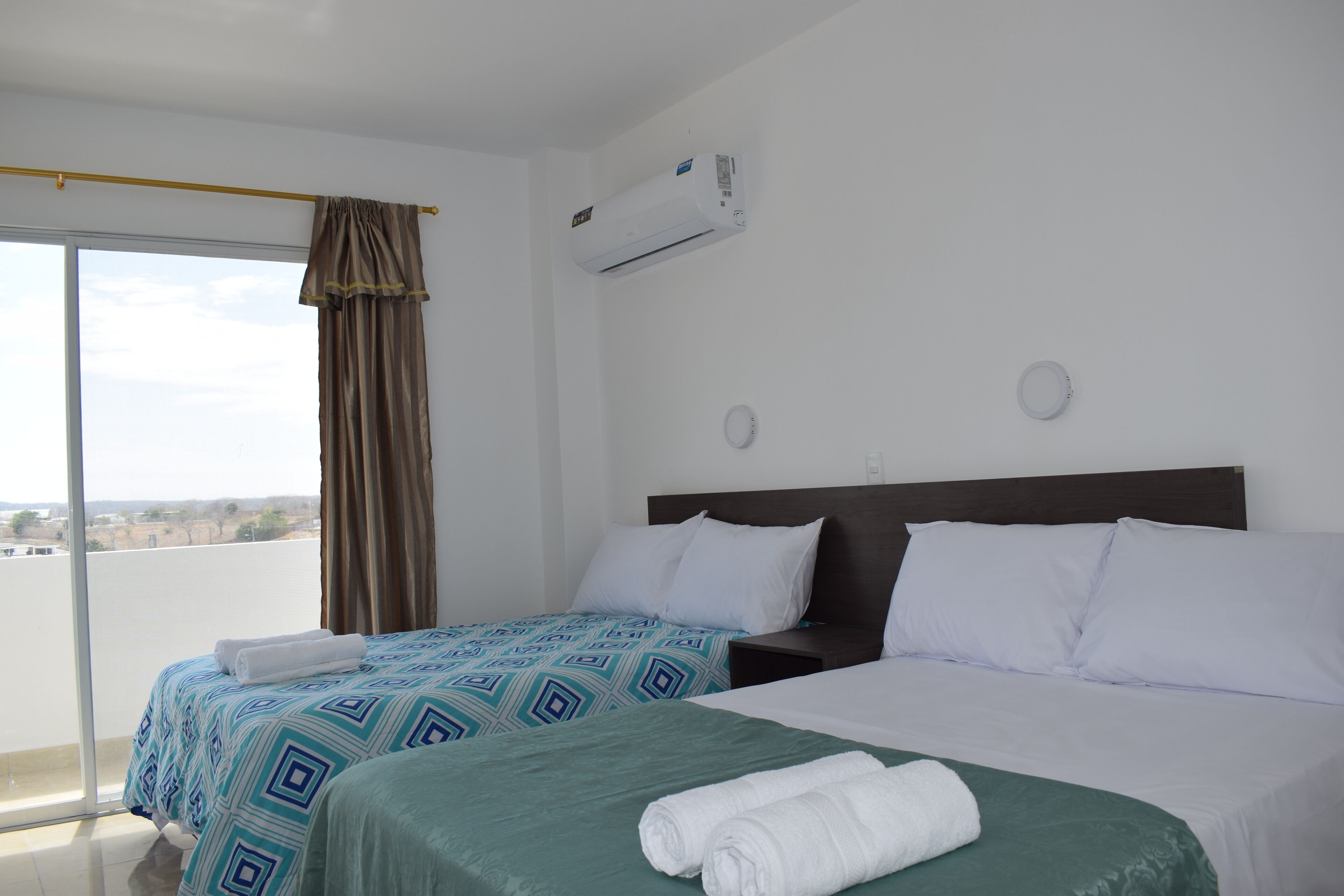 Habitación de hotel con dos camas, un ventanal con cortinas, un equipo de aire acondicionado y vistas a la ciudad.