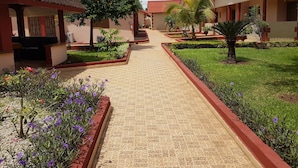 A pathway leading to a row of buildings with red roofs and beige walls, surrounded by well-maintained gardens and a clear sky. - Hôtel La Rose Blanche Korhogo (Korhogo)