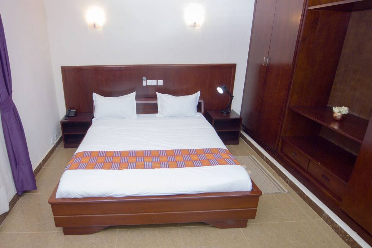 A hotel room with a large bed, two bedside tables, a nightstand, a wooden wardrobe, and a wall-mounted light fixture.
