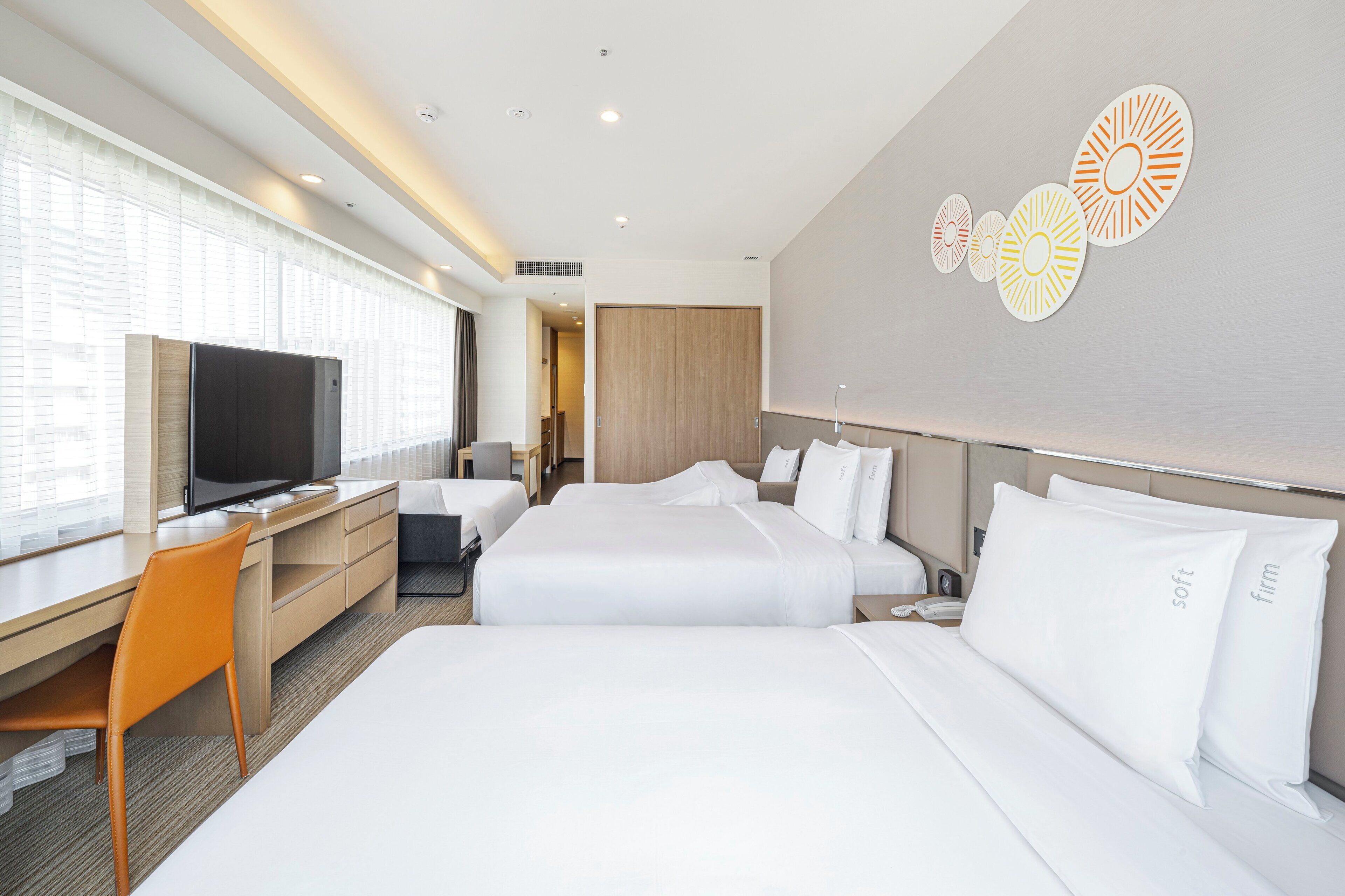 A hotel room with two beds, a desk with a television, and a large window with blinds.