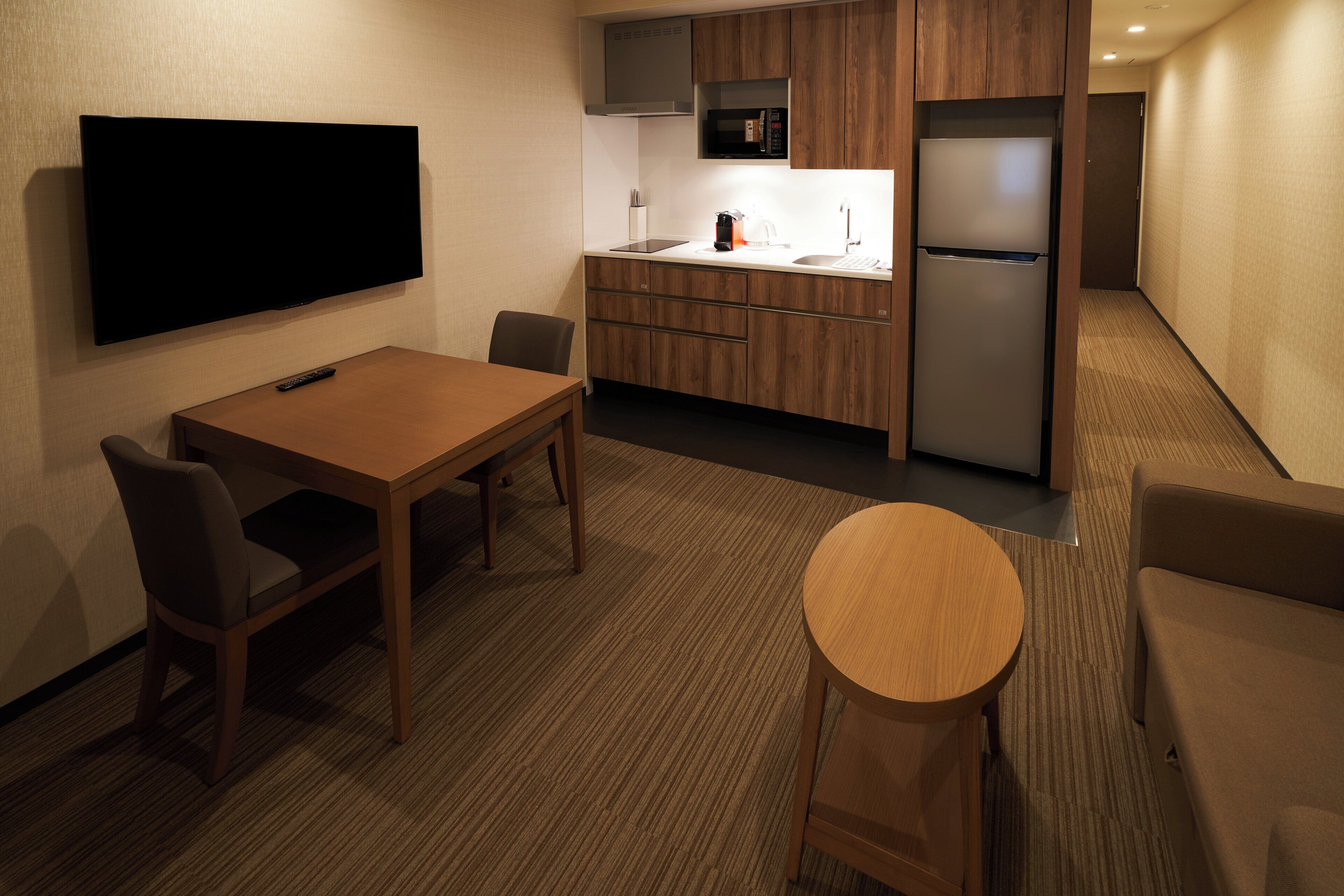 Habitación de hotel compacta con kitchenette, televisor de pantalla plana, mesa de comedor con sillas y una pequeña mesa redonda.
