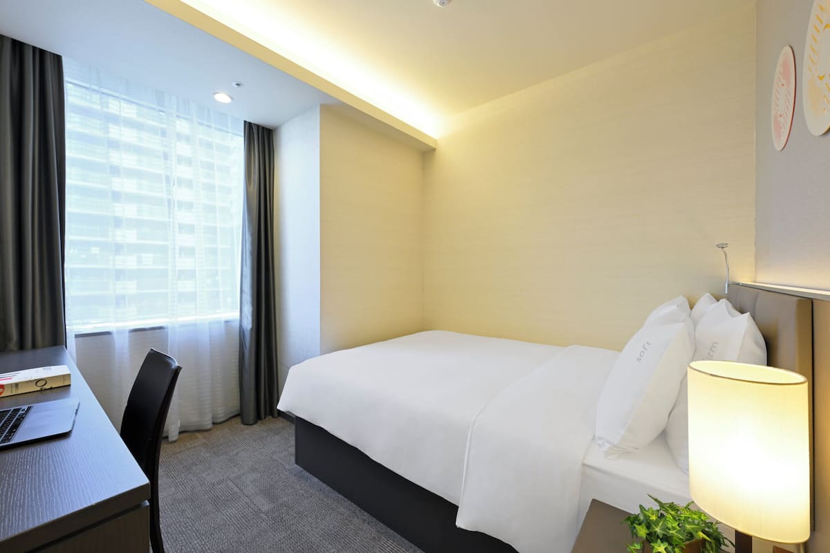 A hotel room with a bed, a desk, a chair, a lamp, and a window with curtains.