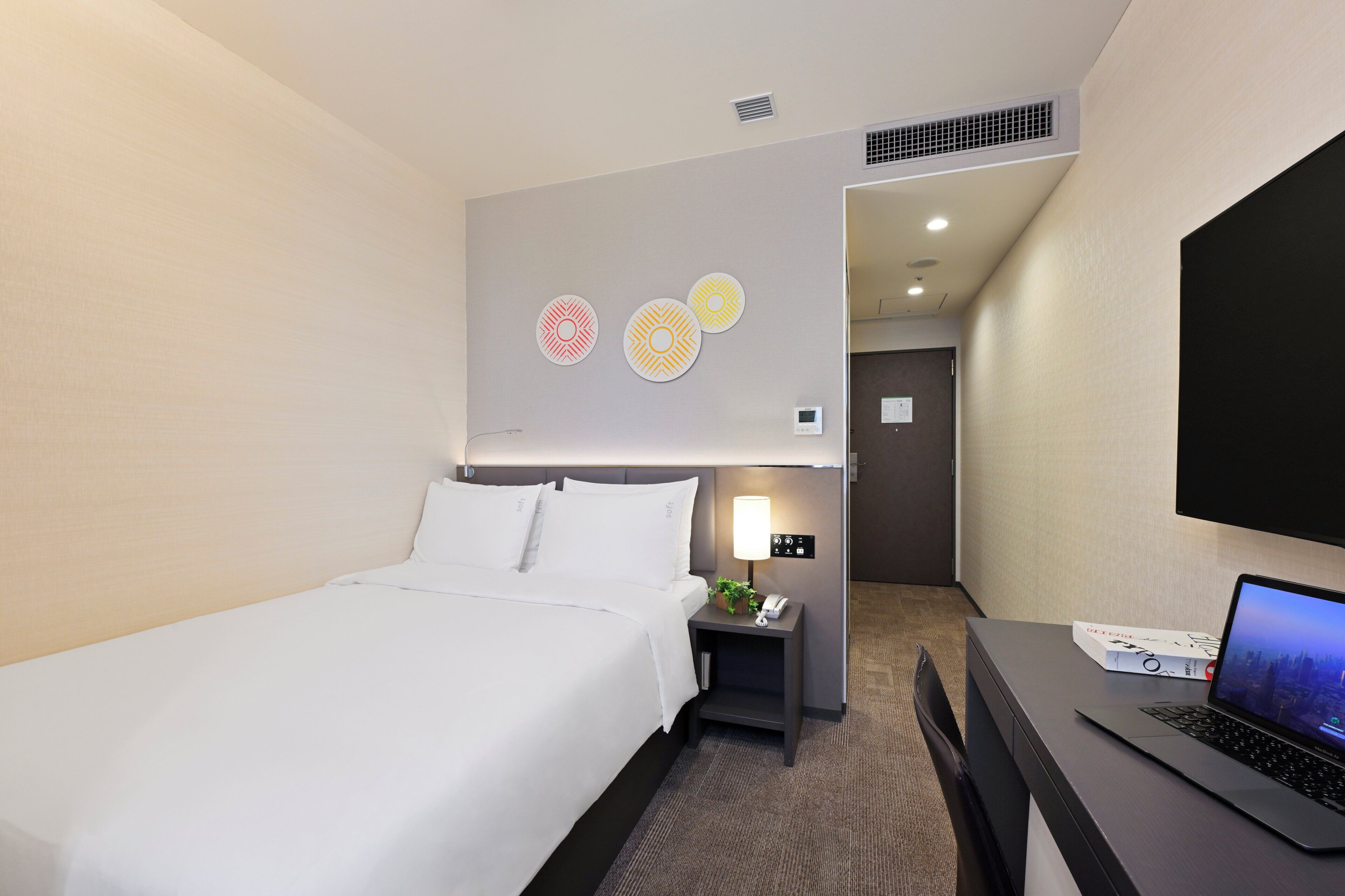 A hotel room with a bed, a desk with a laptop, a nightstand with a lamp, and decorative wall art.