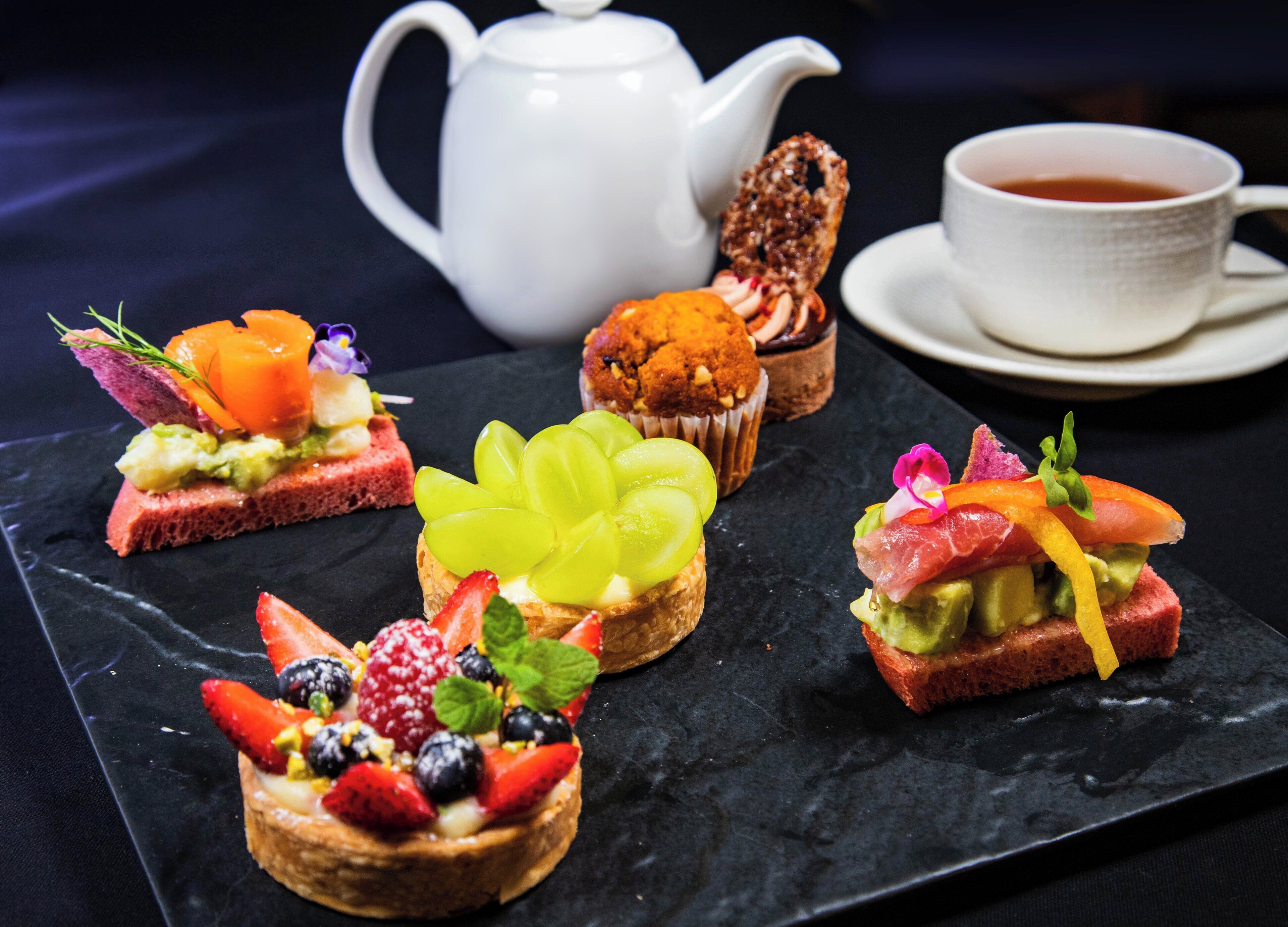 Une sélection de desserts raffinés présentés sur une planche de service en ardoise, une théière blanche et une tasse de thé posée sur une nappe sombre.