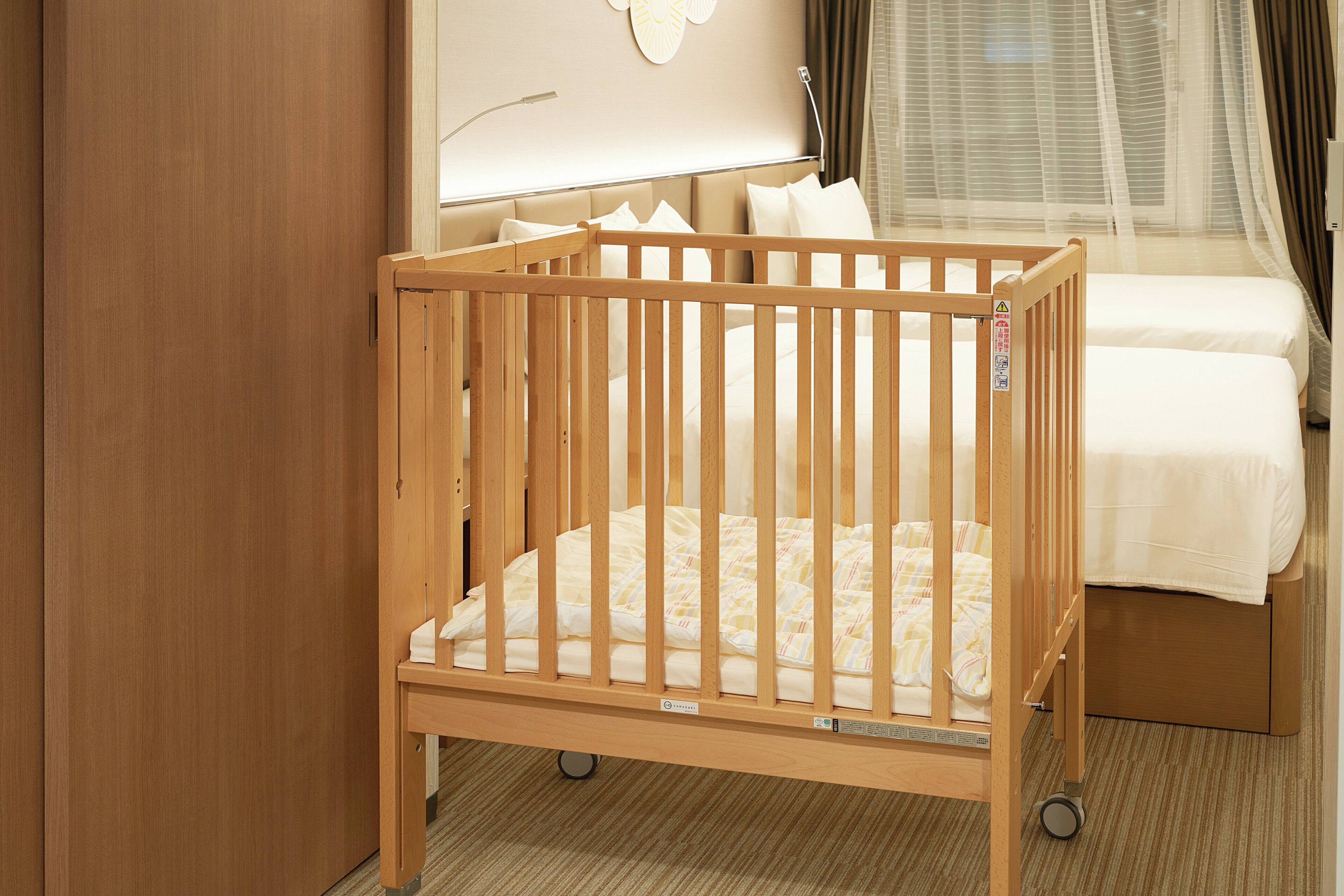 Una cuna de madera con colchón y un cajón pequeño debajo, ubicada en una habitación de hotel con una cama y una ventana con cortinas.