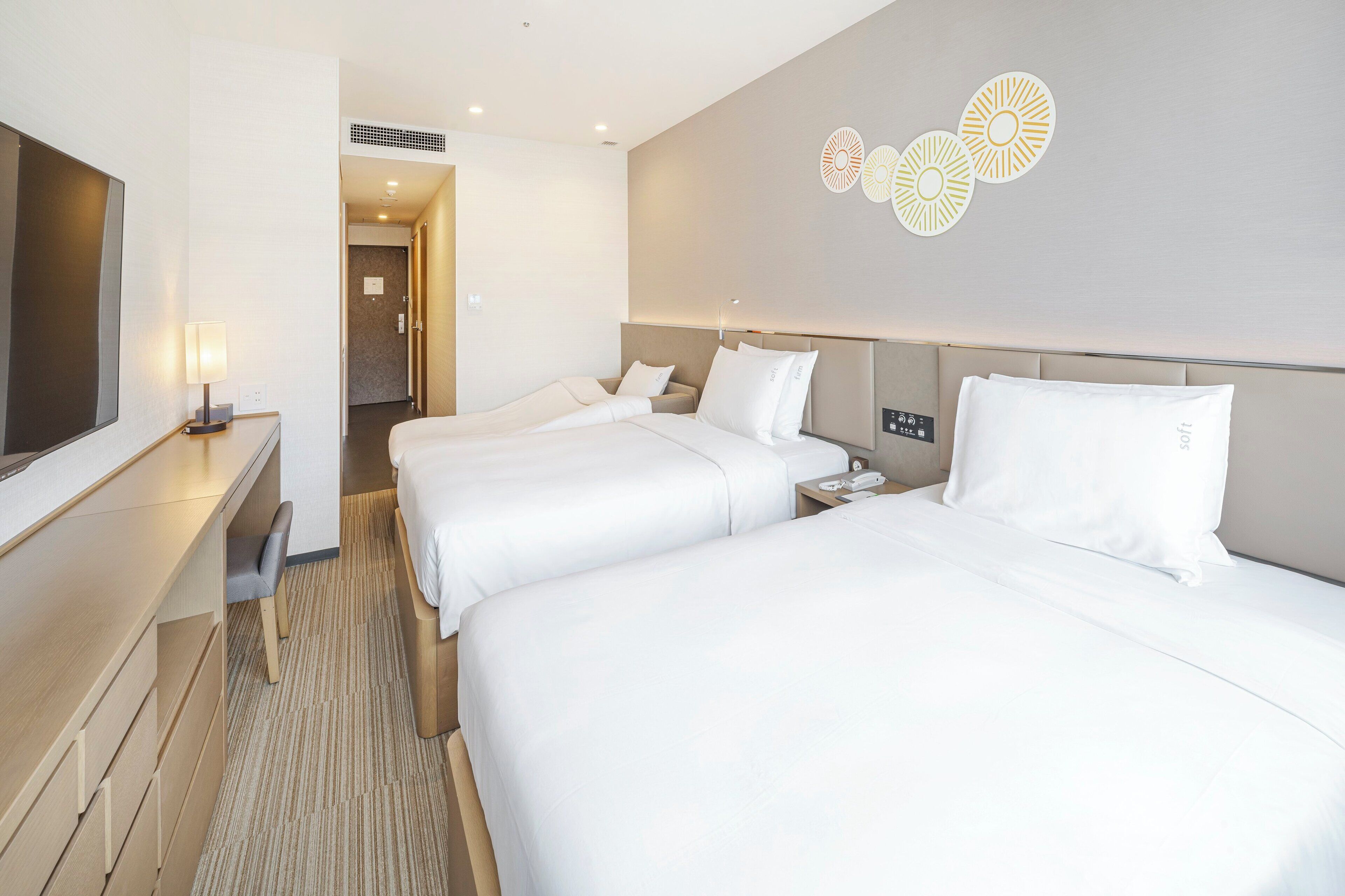 A hotel room with two beds, a desk, a chair, and a wall-mounted TV.