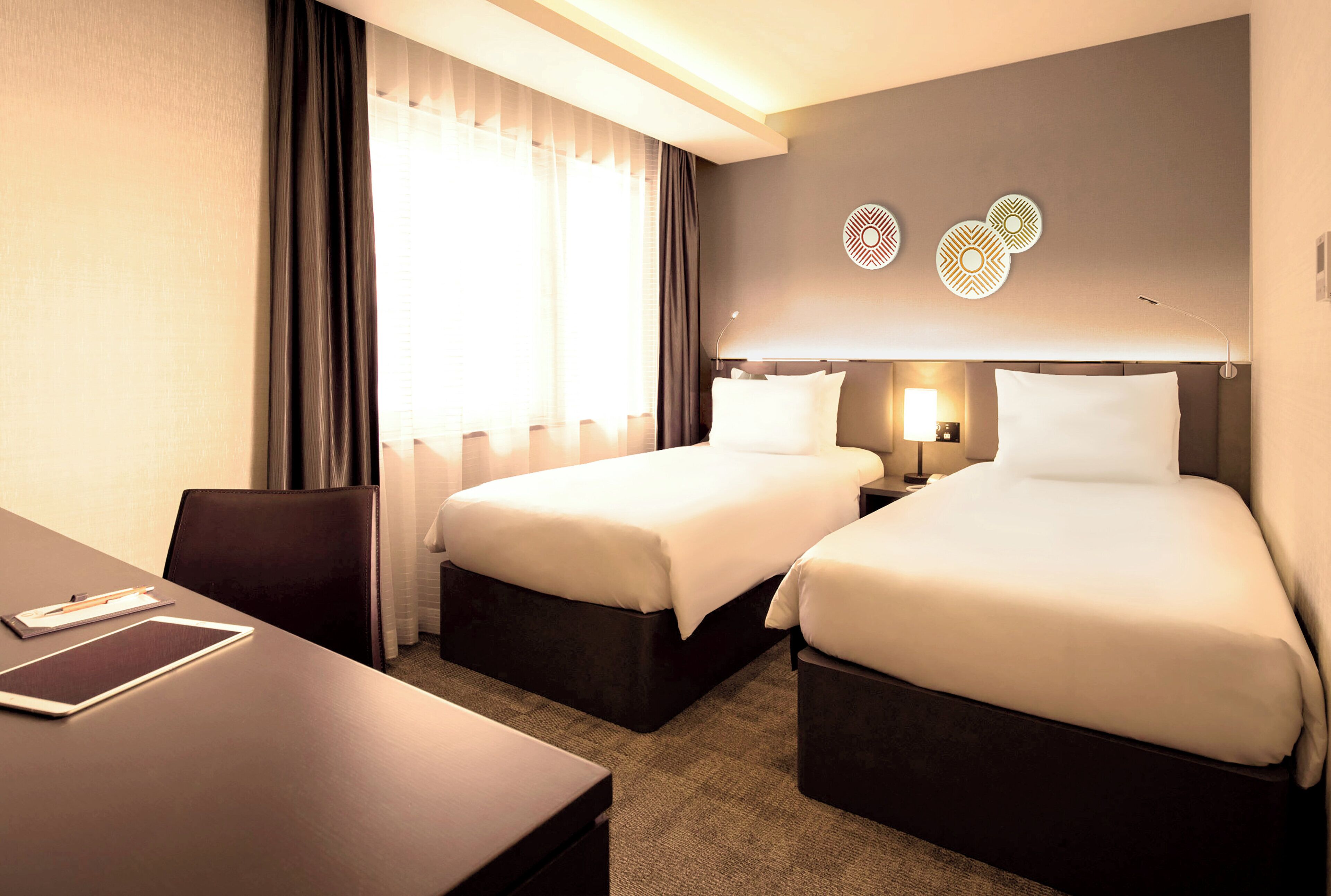 A hotel room with two beds, a desk, and a chair. There are two bedside tables with lamps.