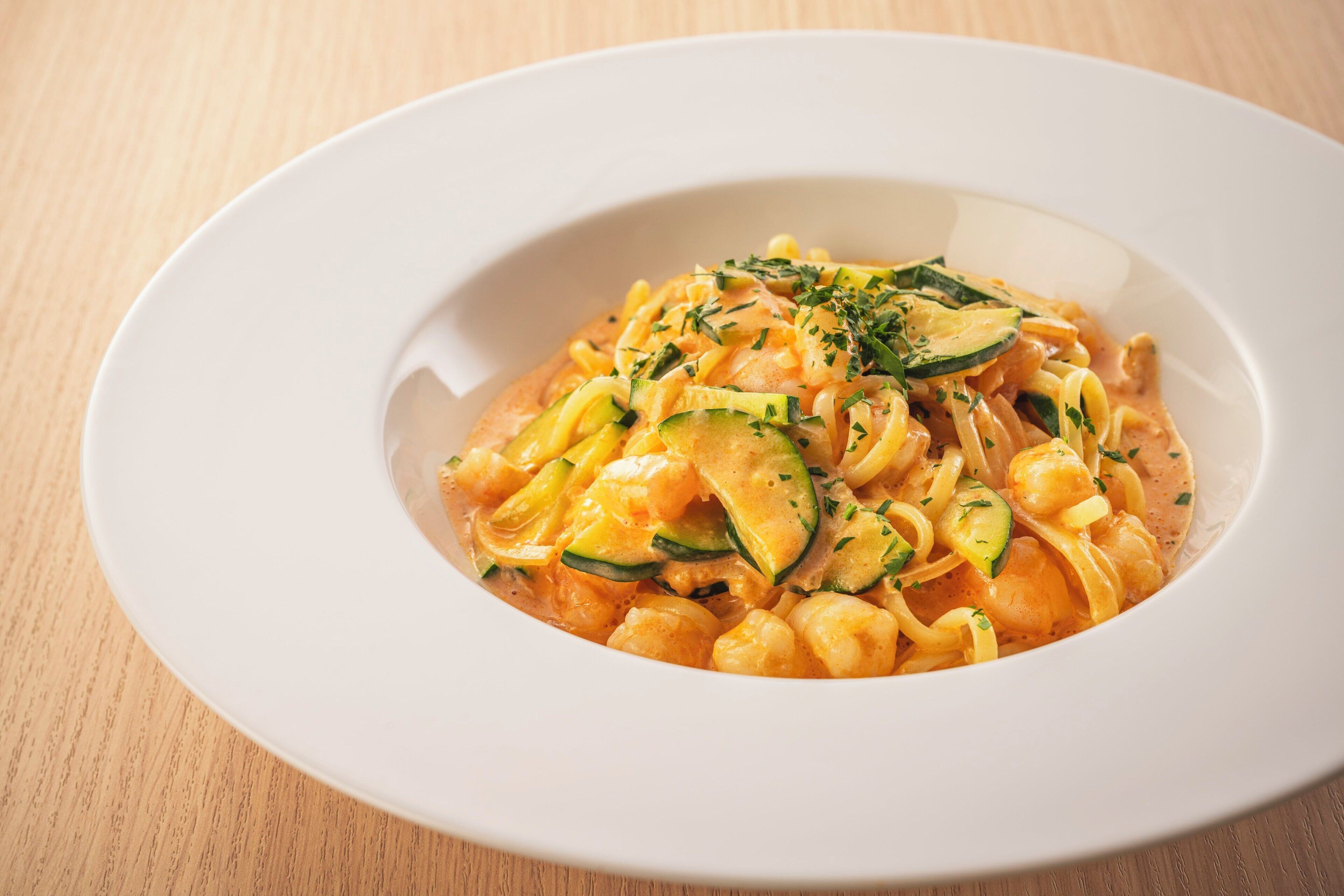 A white plate with pasta, zucchini, and herbs on a wooden table.