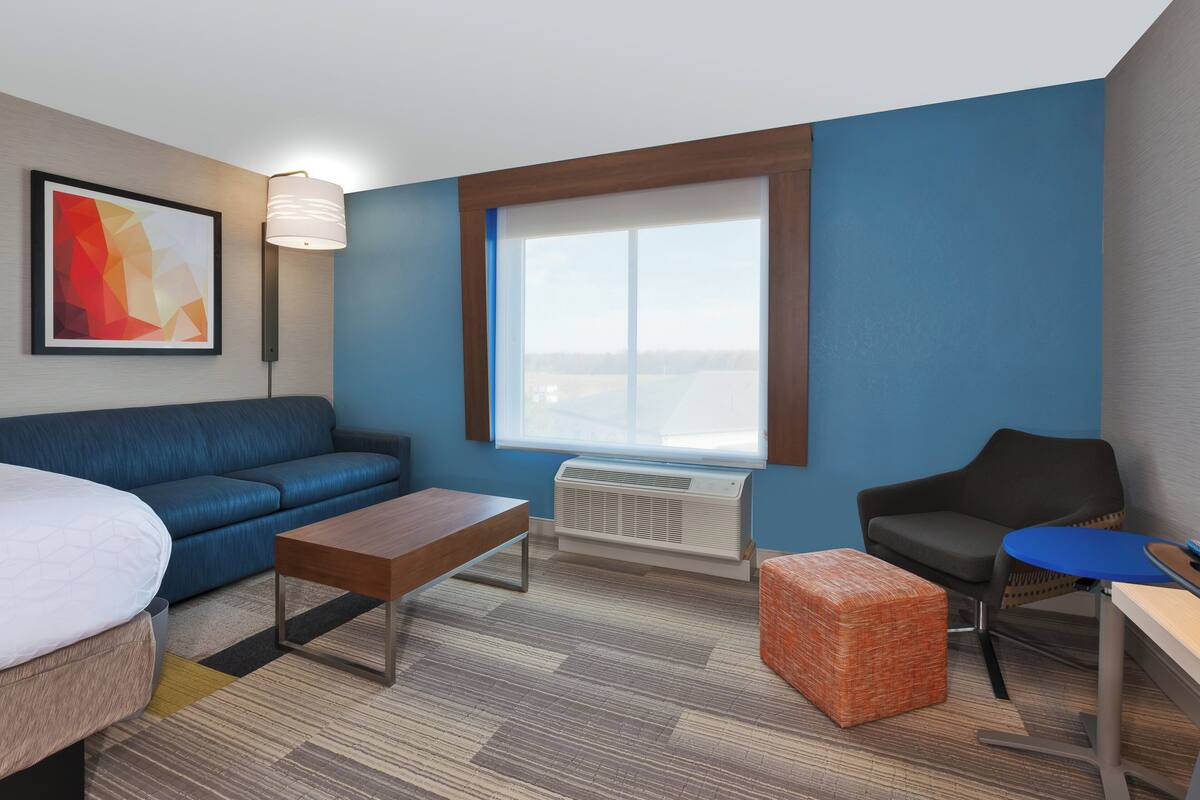 A hotel room with a blue sofa, a wooden coffee table, a small orange ottoman, a desk with a chair, and a large window with a view.