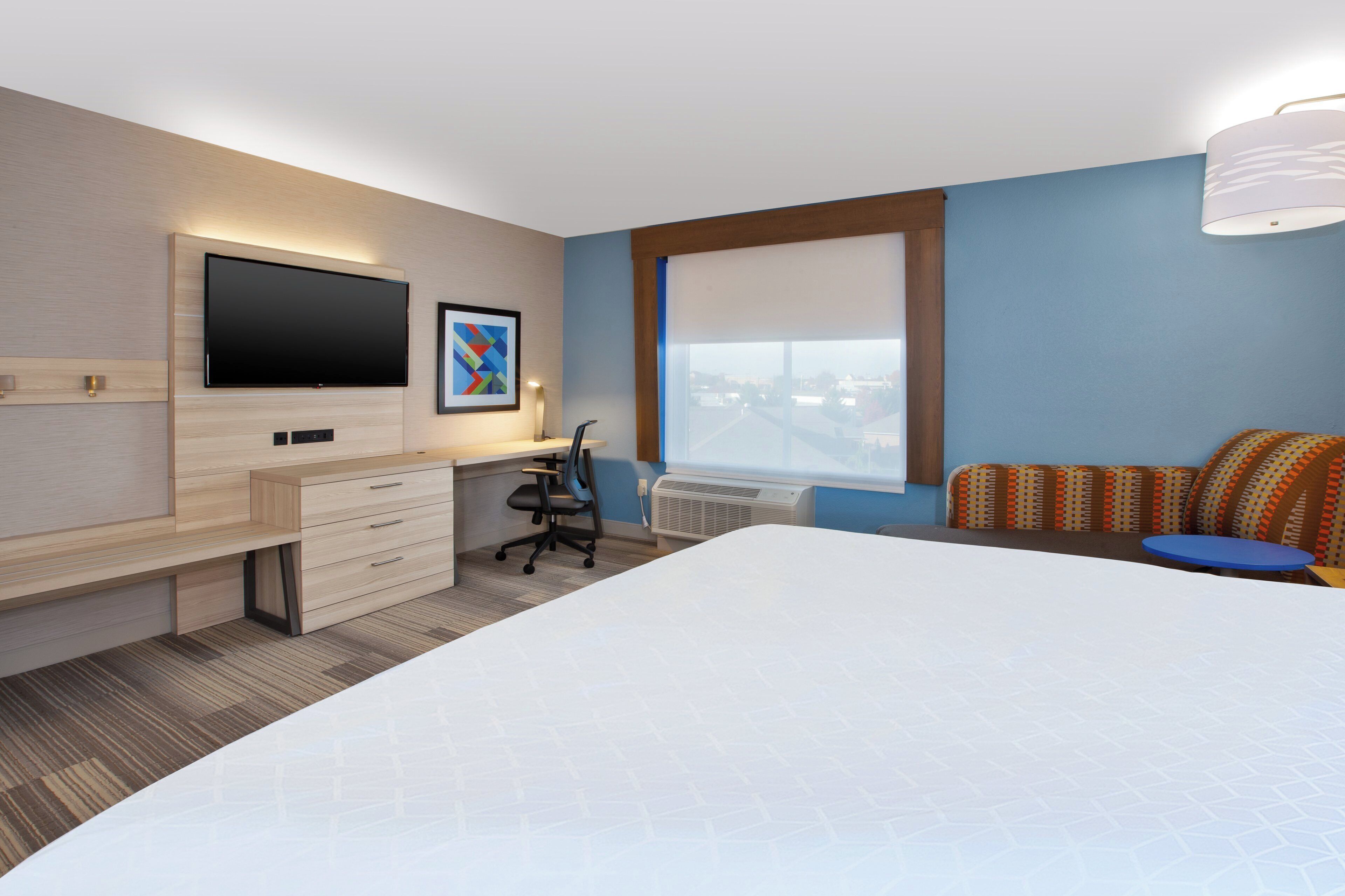 Habitación de hotel con cama, escritorio con silla, televisor de pantalla plana, ventana con vistas y almohada estampada.