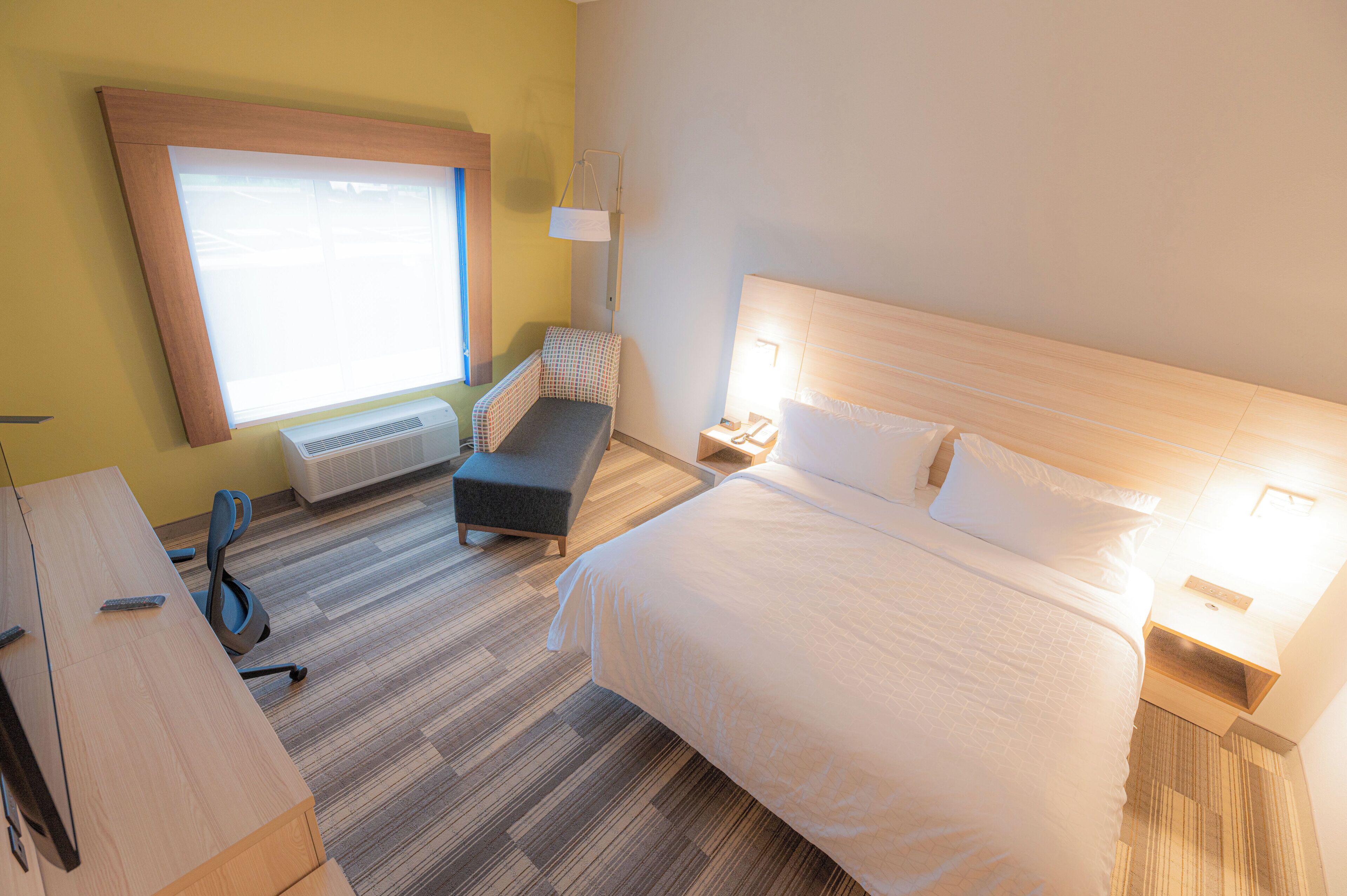 A hotel room with a bed, bedside table, lamp, and a window with blinds.