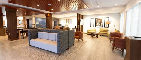 A spacious lobby with a modern design, featuring a central seating area with a sofa and chairs, a reception desk, and a dining area.