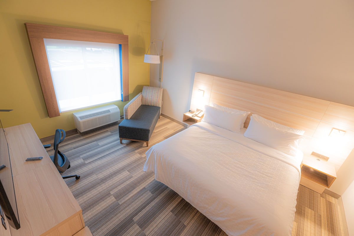 Ein Hotelzimmer mit einem großen Bett, einem Schreibtisch, einem Sessel, einem Nachttisch mit Lampe und einem Fenster mit Jalousien.