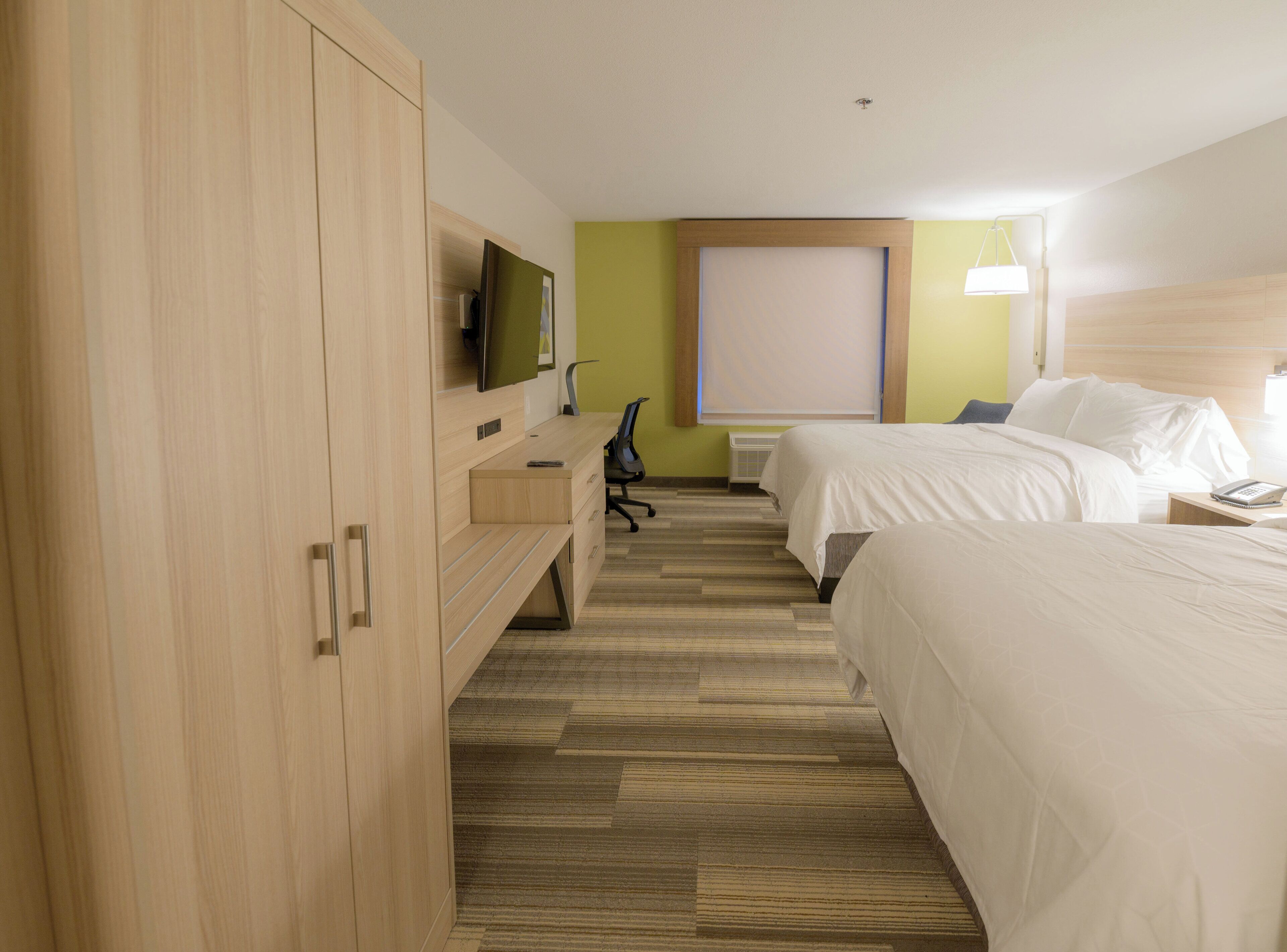 Une chambre d’hôtel avec une armoire en bois, un bureau avec une chaise, deux lits, un téléviseur et un climatiseur fixé au mur.