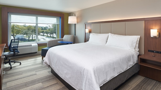 A modern hotel room with a large bed, a desk, and a chair. There is a window with a view of trees and buildings. - Holiday Inn Express Doral Miami by IHG (Miami)