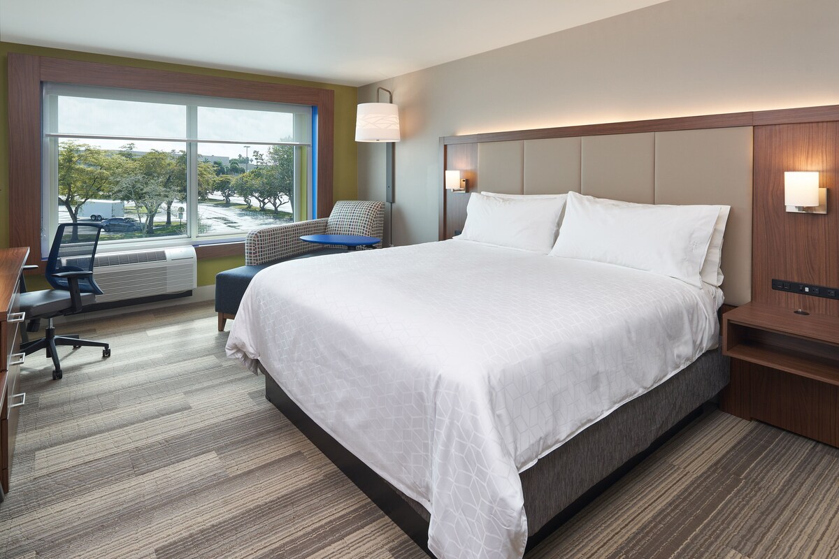 A modern hotel room with a large bed, a desk, and a chair. There is a window with a view of trees and buildings.