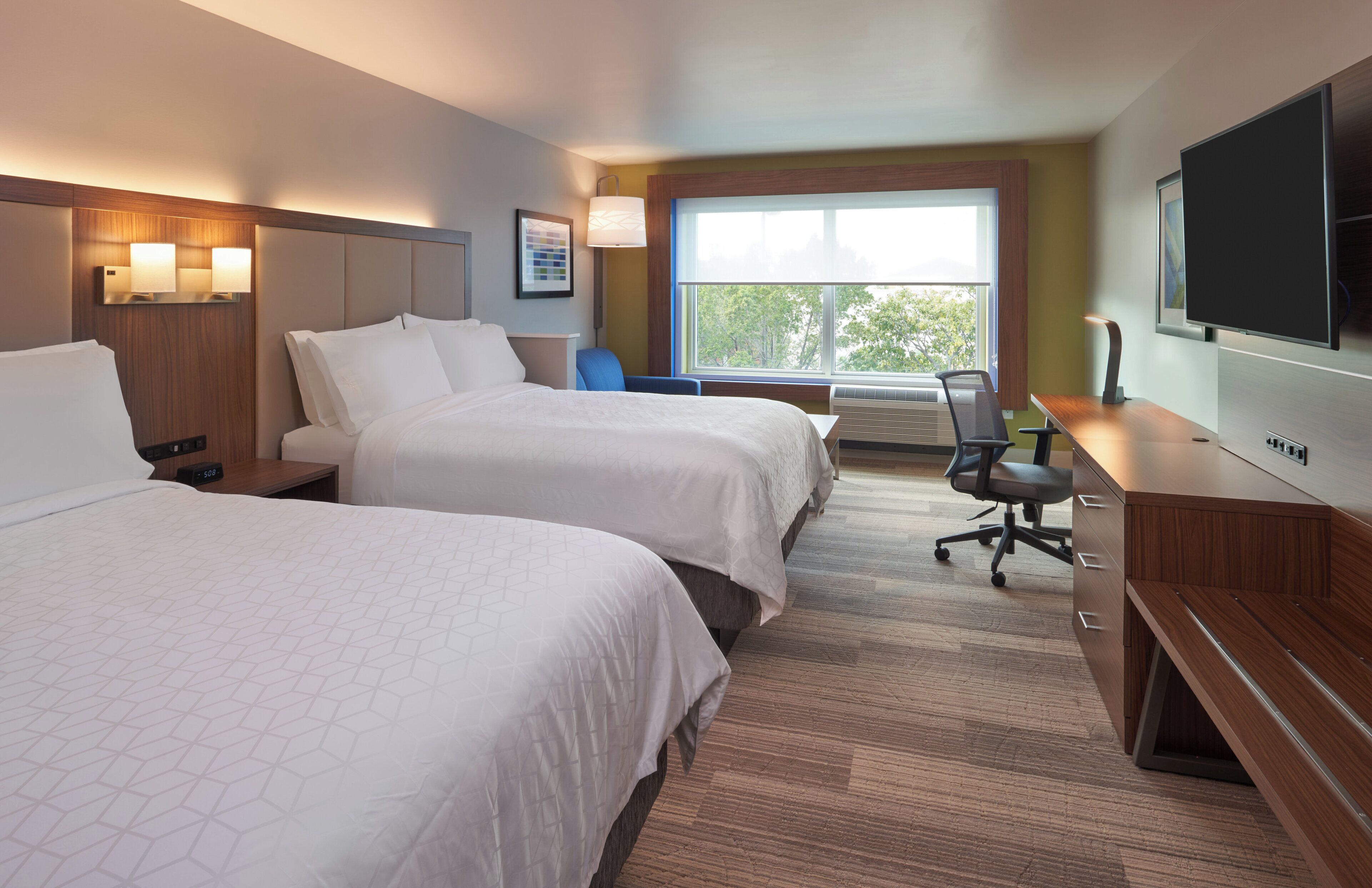 A hotel room with two beds, a desk, a chair, a TV, and a window with a view of trees.