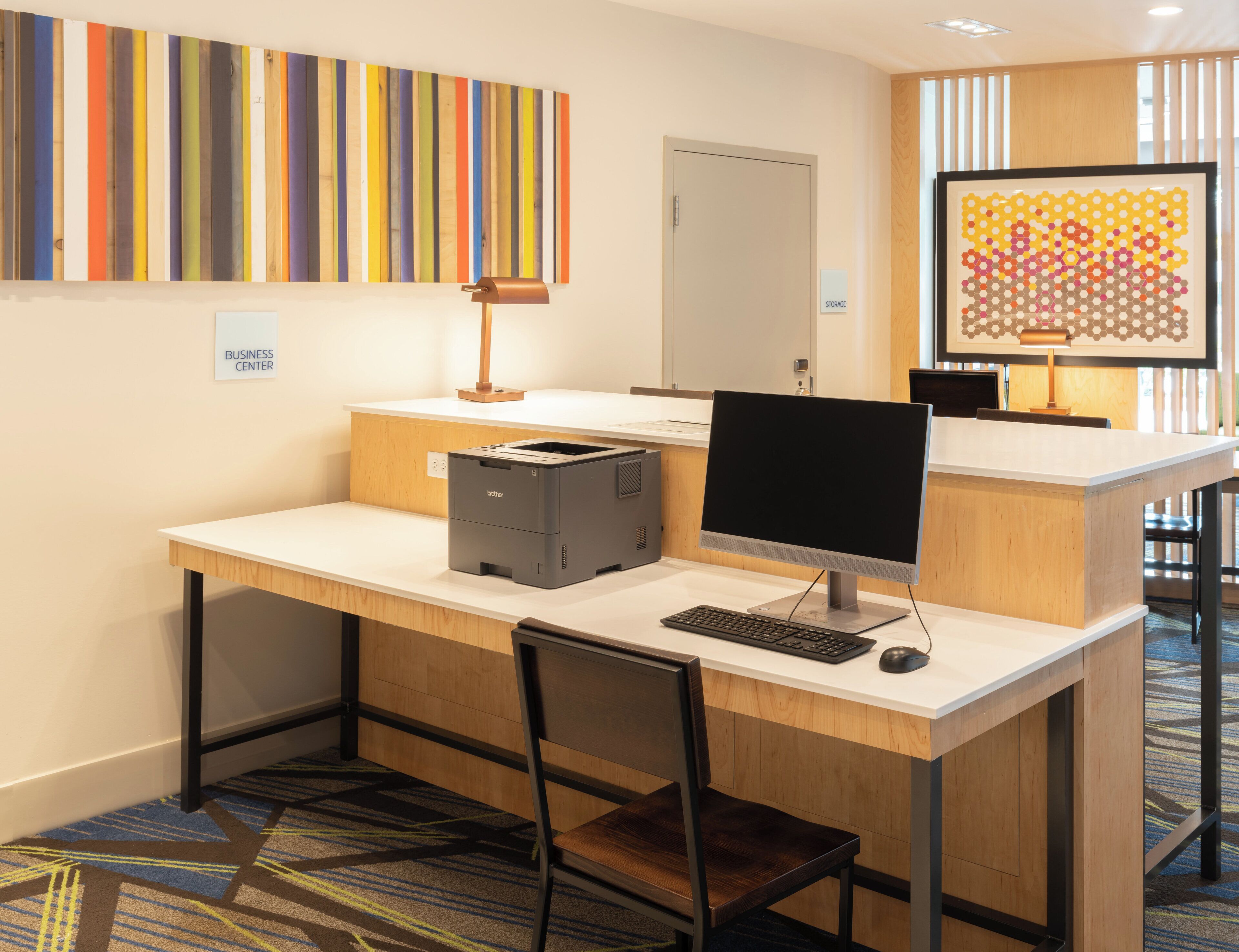 A hotel business center with a desk, computer, printer, and a colorful abstract painting on the wall.