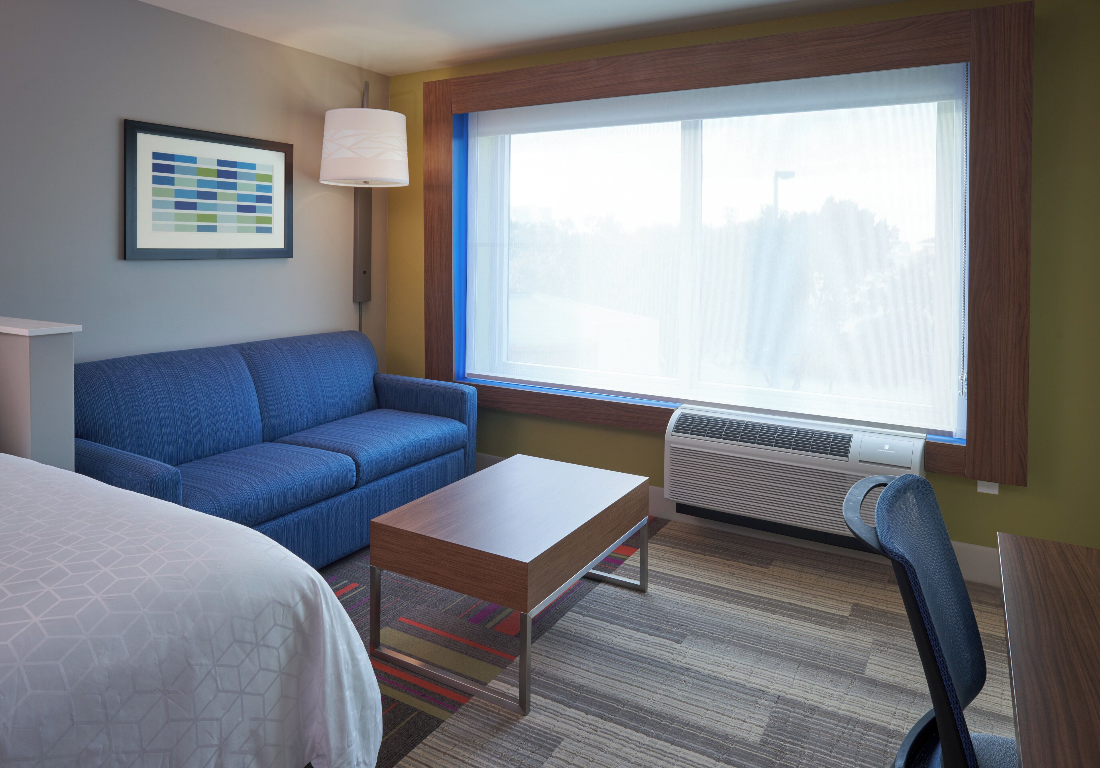 A hotel room with a blue sofa, a small wooden table, a single bed, a large window with a view, and a wall-mounted air conditioner.