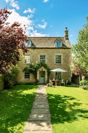 A stone house with a green lawn, a pathway leading to the entrance, and a table with an umbrella.