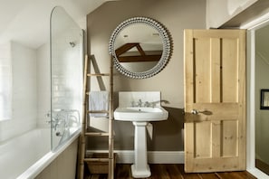 A bathroom with a glass-enclosed shower, a wooden door, a white pedestal sink, and a mirror with a decorative frame.