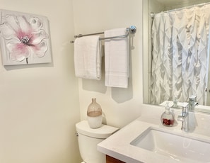 A bathroom with a toilet, two white towels hanging on a rack, a large mirror, a shower curtain, and a sink with a soap dispenser. - Hollywood Studios in Downtown Phoenix (Phoenix)