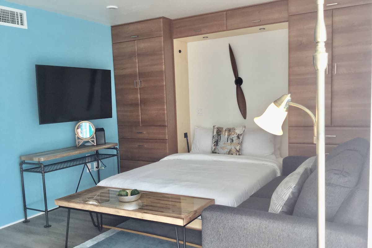 Une chambre moderne avec un lit, un canapé gris, un bureau en bois avec un téléviseur à écran plat et une lampe de chevet.