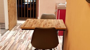 Un coin repas avec une table et des chaises en bois, une cuisine rouge et blanche en arrière-plan, et un balcon avec une balustrade.