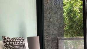 A bedroom with a bed, a patterned pillow, a lamp, and a view of a brick wall and trees outside. - LUXURY APARTMENT IN KAAN HA, BAHIA PRINCIPE (Akumal)