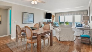 Un coin repas avec une table et des chaises en bois, un ventilateur de plafond et une vue sur l’océan grâce à de grandes fenêtres.