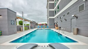 Una piscina con un letrero que dice 'NO DIVING', rodeada por un edificio moderno de paredes grises y un área techada con sillas amarillas.