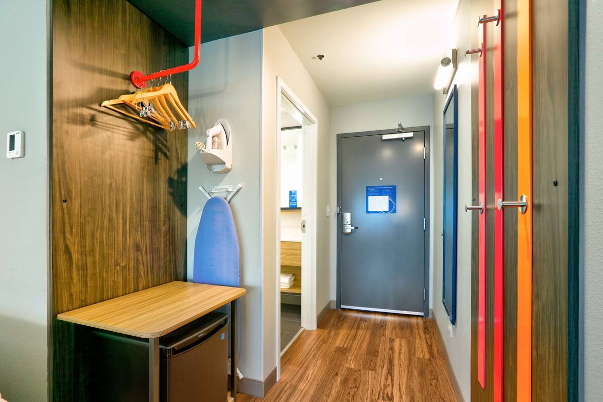 A hotel corridor with a wooden bench, a compact refrigerator, a wall-mounted ironing board, and a door with a blue informational sign.