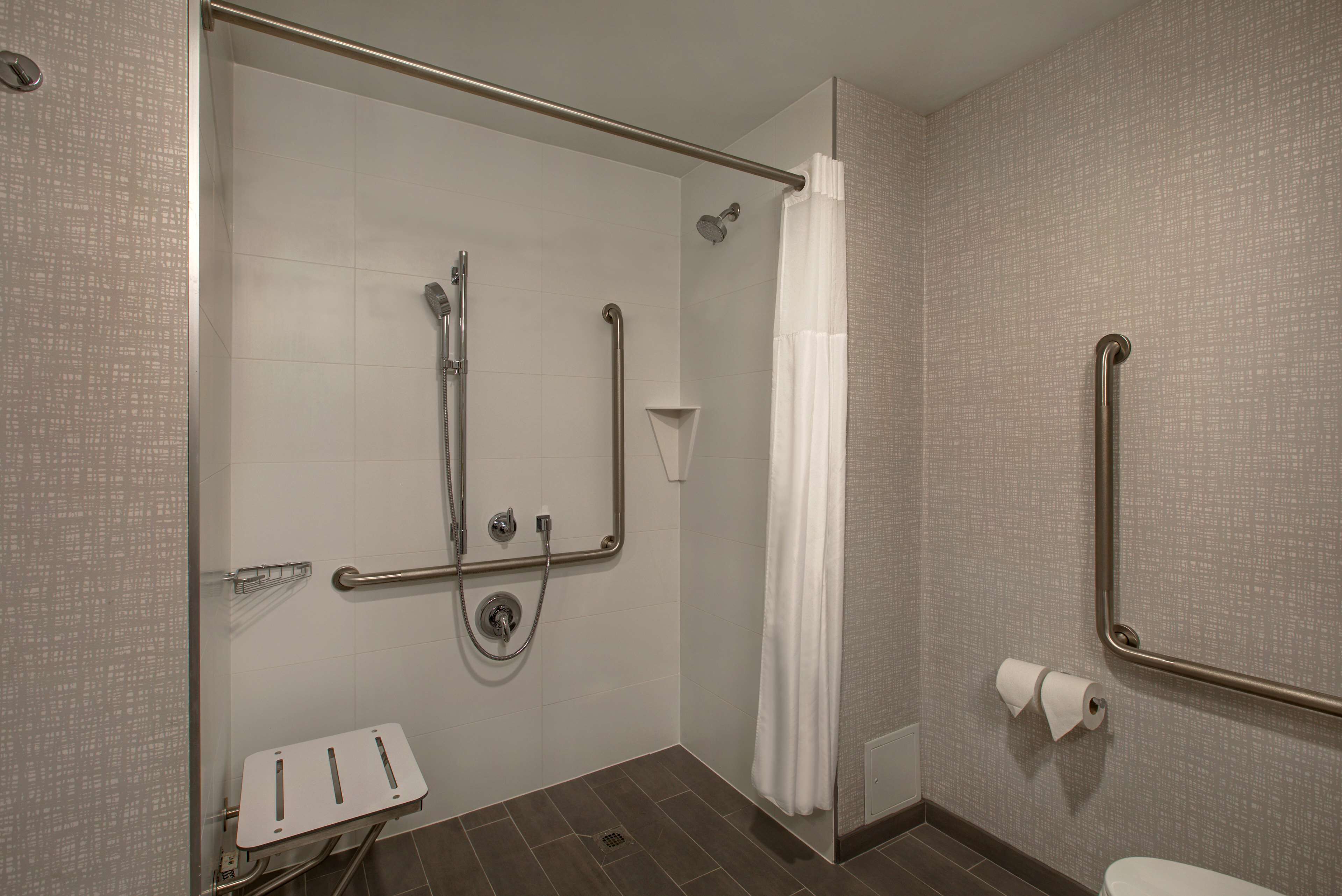 A handicap-accessible bathroom with a shower, grab bars, a toilet, and a white towel dispenser.