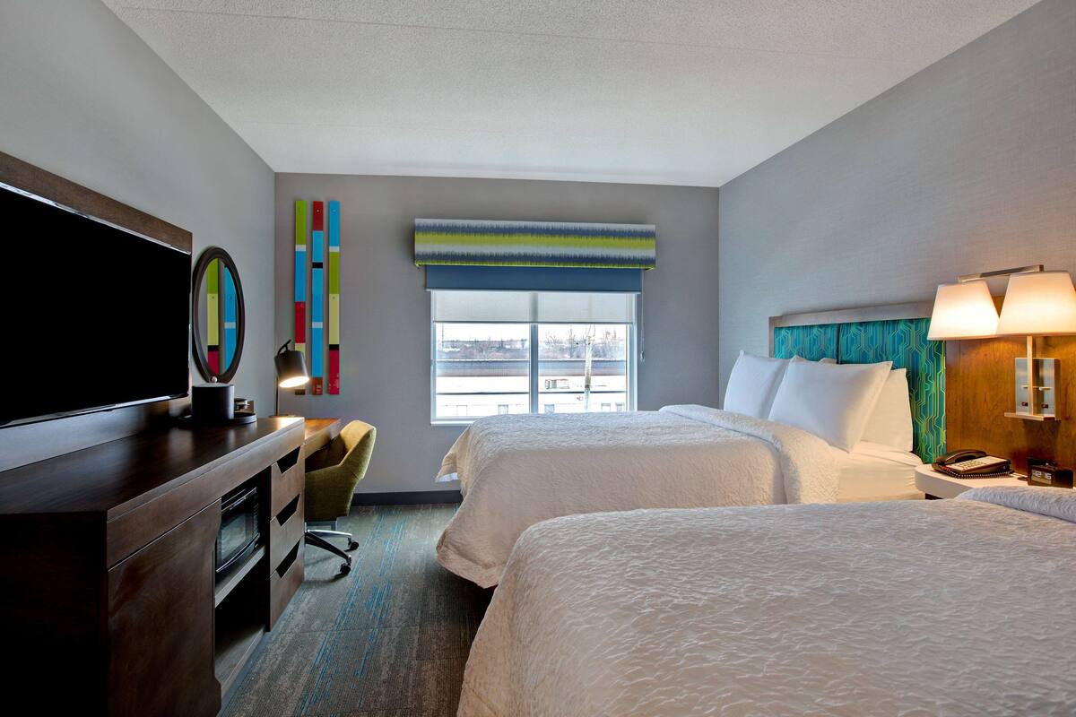 Ein Hotelzimmer mit zwei Betten, einem Schreibtisch, einem Flachbildfernseher, einem Sessel, einem Fenster mit Aussicht und einer Lampe.