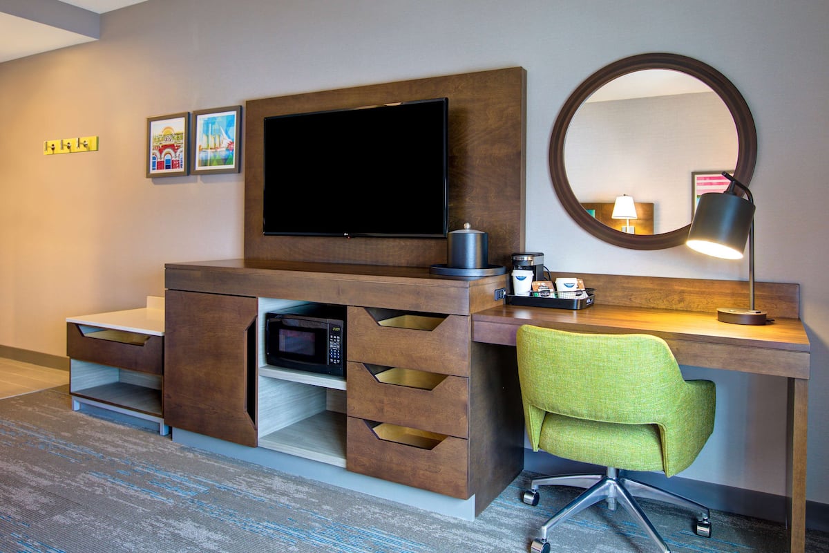 Ein Hotelzimmer mit Schreibtisch aus Holz, einem grünen Sessel, einem Flachbildfernseher, einer Kaffeemaschine und einem runden Spiegel.