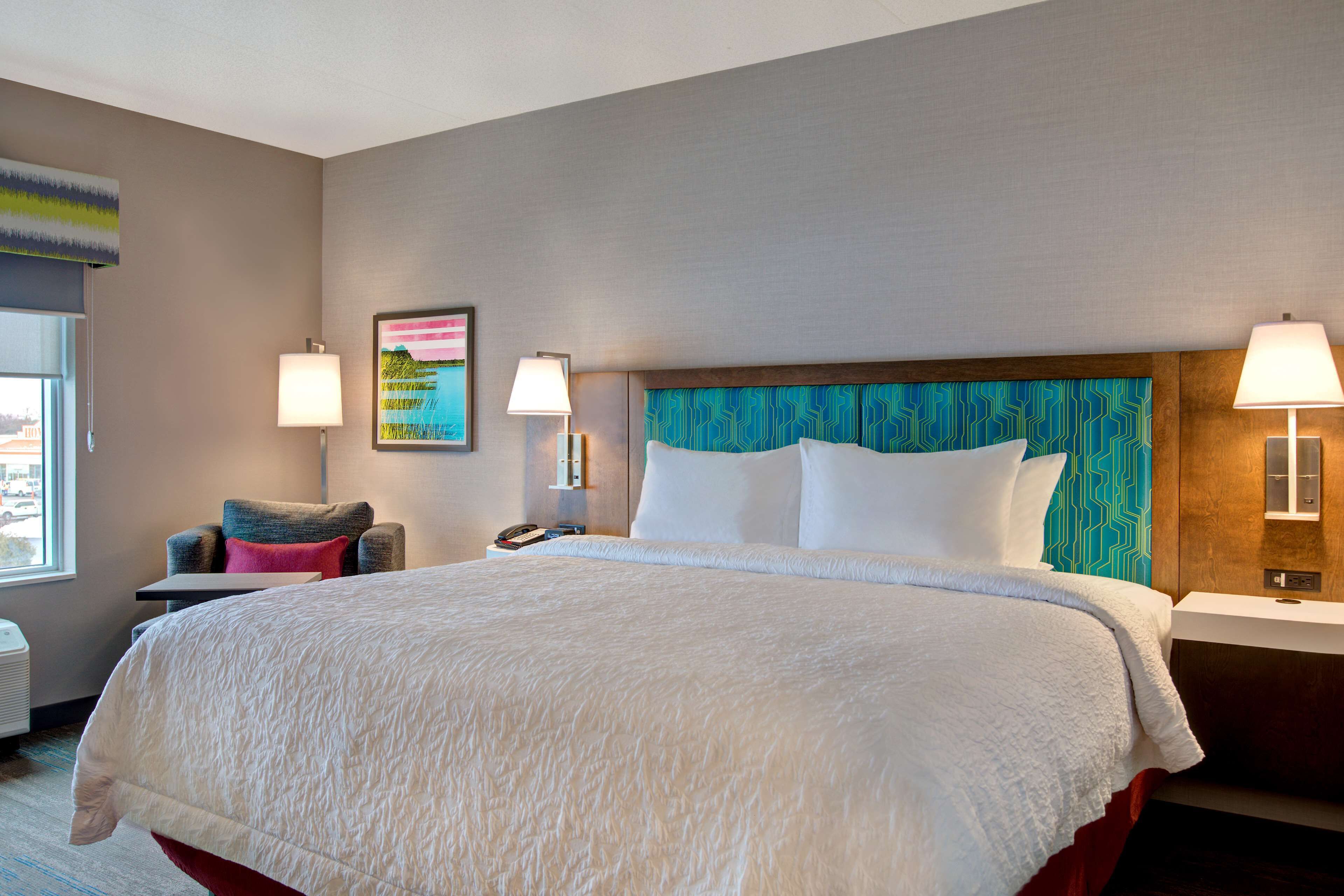 A hotel room with a large bed, a chair, a nightstand with a lamp, and a wall with a painting.