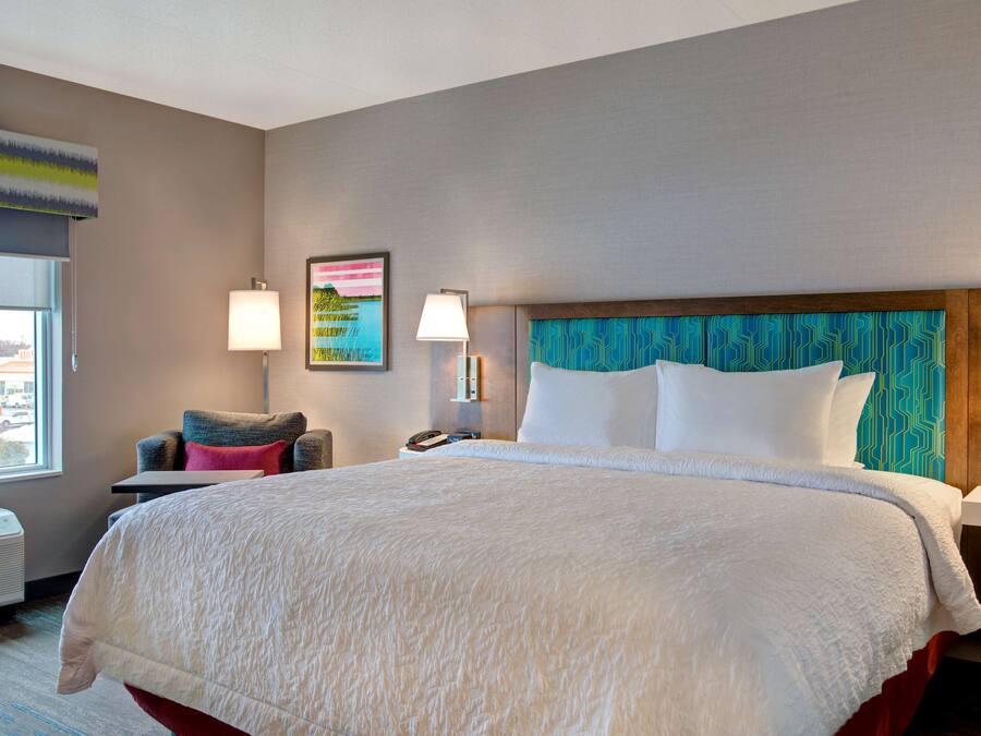 A hotel room with a large bed, a chair, a nightstand with a lamp, and a wall with a painting.