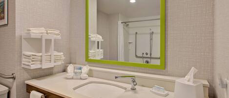A bathroom with a large mirror, a sink, a shower, and neatly folded towels on a shelf.