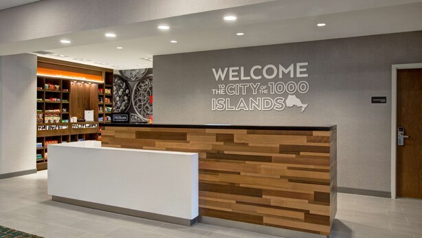 A reception desk with a "Welcome to the City of 1000 Islands" sign, a wooden reception area, and a background with a wall-mounted map and shelves with various items. - Hampton Inn by Hilton Brockville (Brockville)