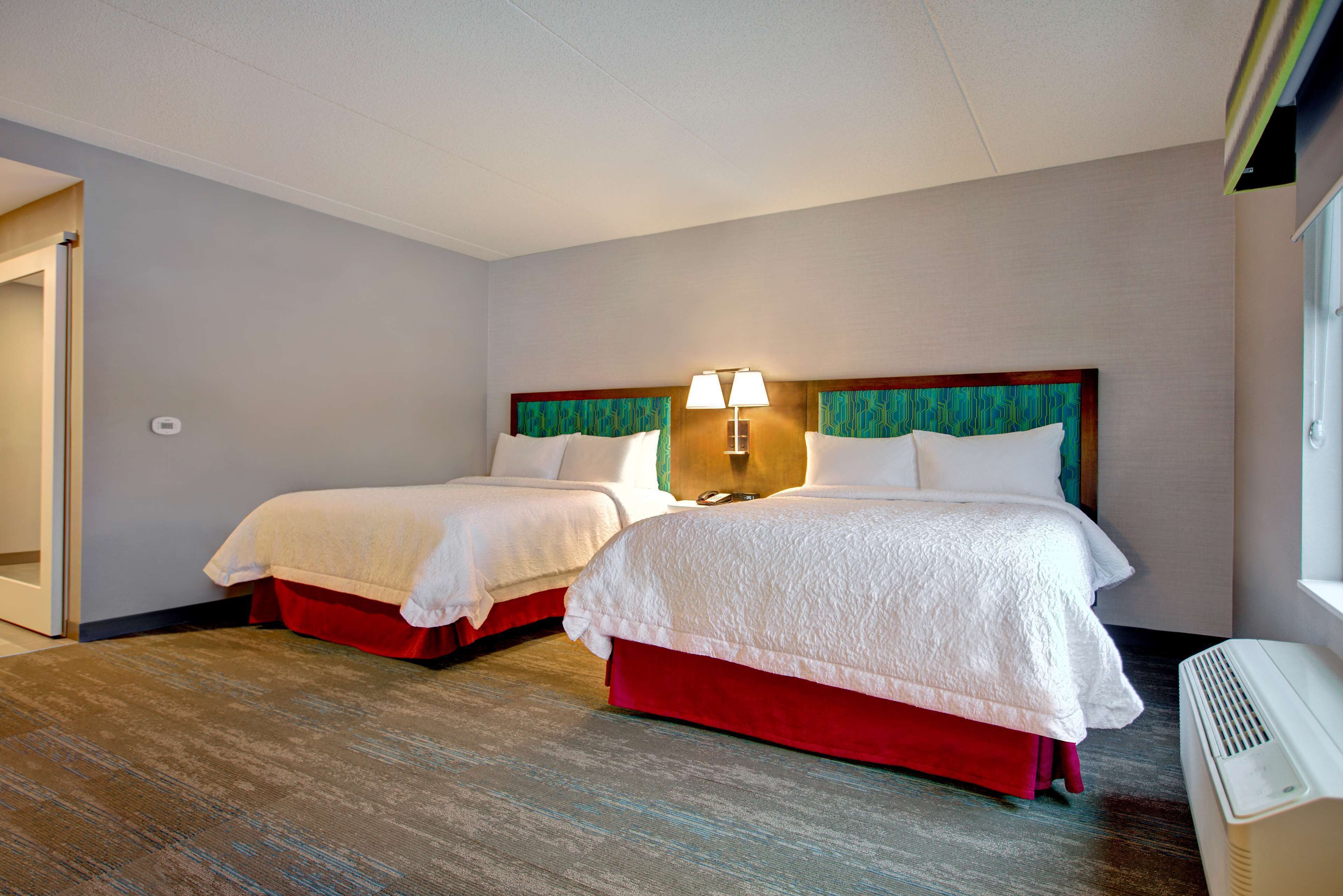 A hotel room with two beds, a wall-mounted lamp, and a window.