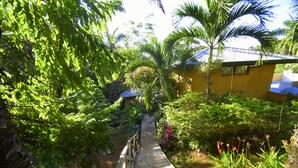 A wooden pathway leading through a lush garden with various plants and a yellow building in the background. - Villas Punta India (Cuajiniquil)