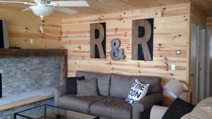 A cozy living room with a wooden ceiling, a ceiling fan, a stone fireplace, and a couch with decorative pillows. - Lake Front Cottage Right Across Street ORV And Snowmobile, Blue Bear trail (Mancelona)