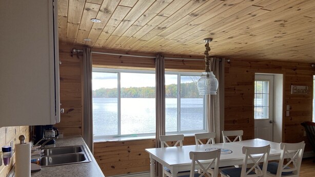A cabin with a kitchen, dining area, and large windows overlooking a lake. - Lake Front Cottage Right Across Street ORV And Snowmobile, Blue Bear trail (Mancelona)