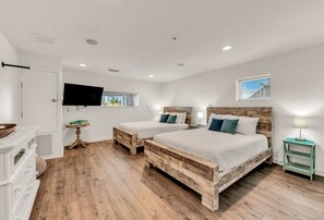 Une chambre avec deux lits en bois, un téléviseur fixé au mur, une fenêtre avec des stores et une petite table avec une lampe.