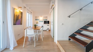 Un interior moderno con una mesa de comedor de madera, sillas blancas, una zona de cocina con televisor y una escalera con escalones de madera.