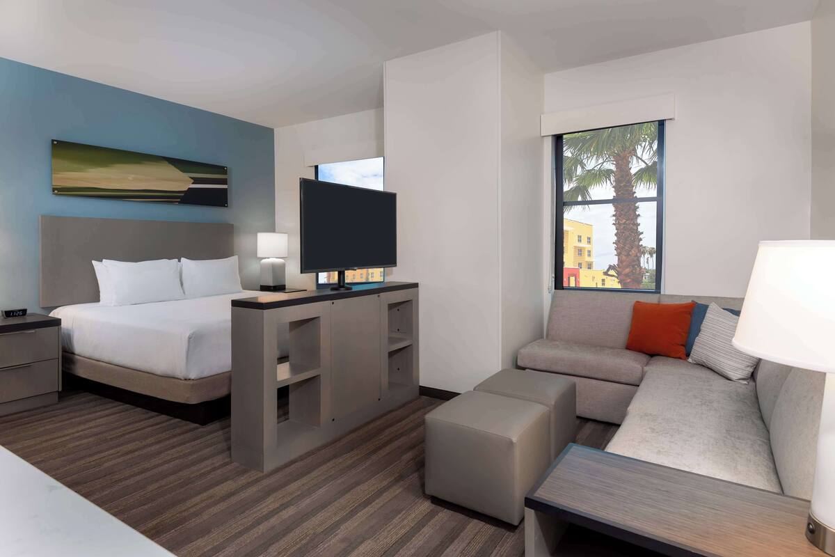 A modern hotel room with a bed, a desk with a flat-screen TV, a sofa, and a window with a view of palm trees.