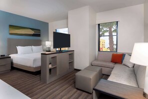 A modern hotel room with a bed, a desk with a flat-screen TV, a sofa, and a window with a view of palm trees. - Hyatt House Tampa Airport Westshore (Tampa)