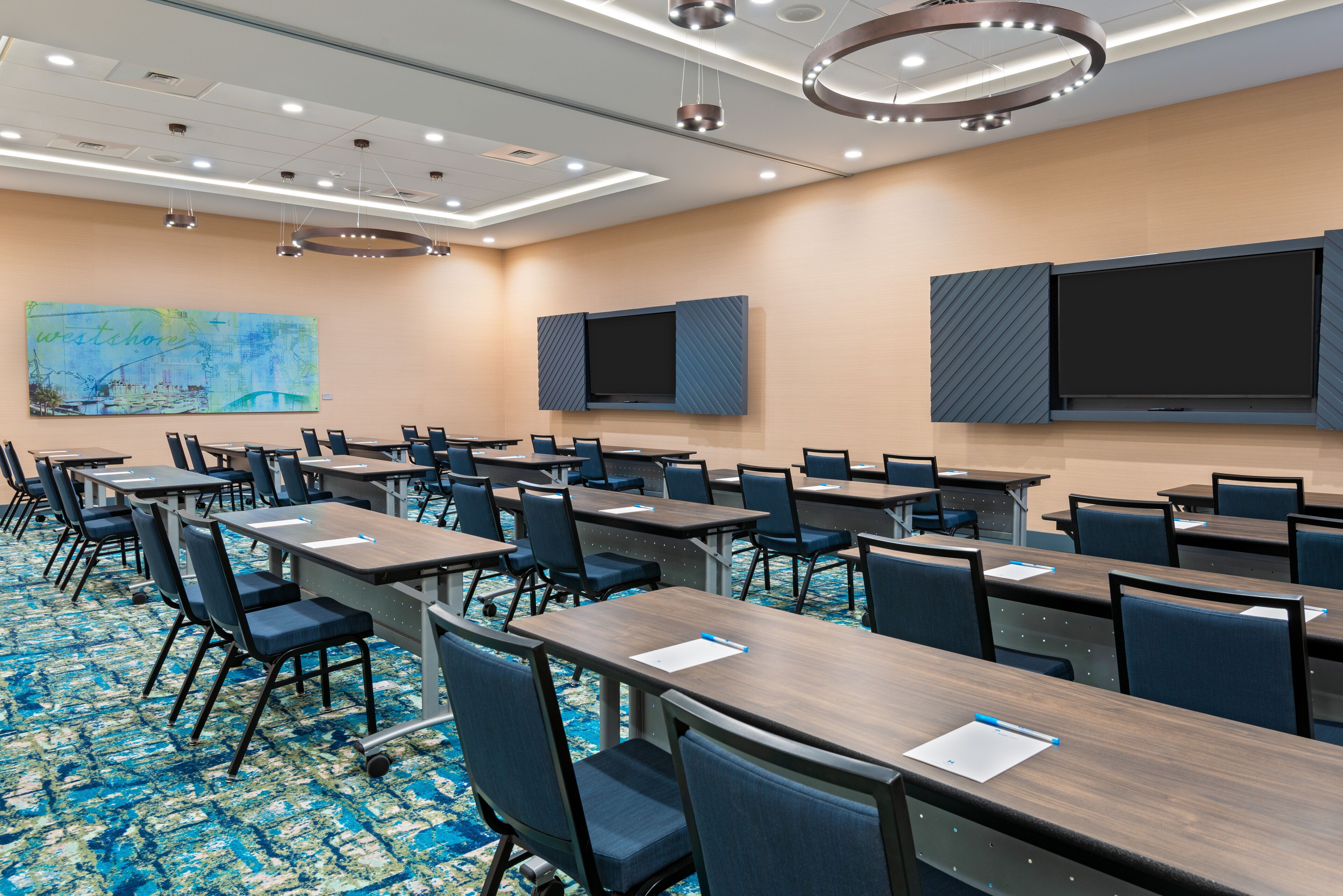 A conference room with multiple tables and chairs, a large flat-screen TV mounted on the wall, and a ceiling with recessed lighting.
